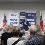 Speaker in a blue suit addresses an indoor political rally, with Roy Cooper banners and American flags behind him.