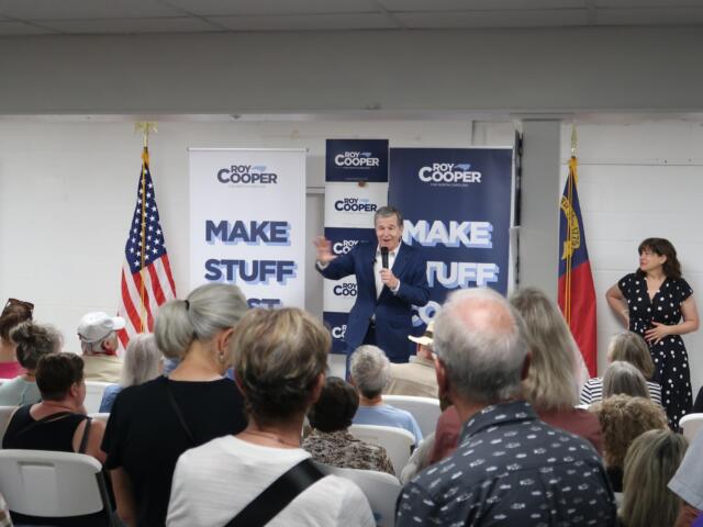 Speaker in a blue suit addresses an indoor political rally, with Roy Cooper banners and American flags behind him.