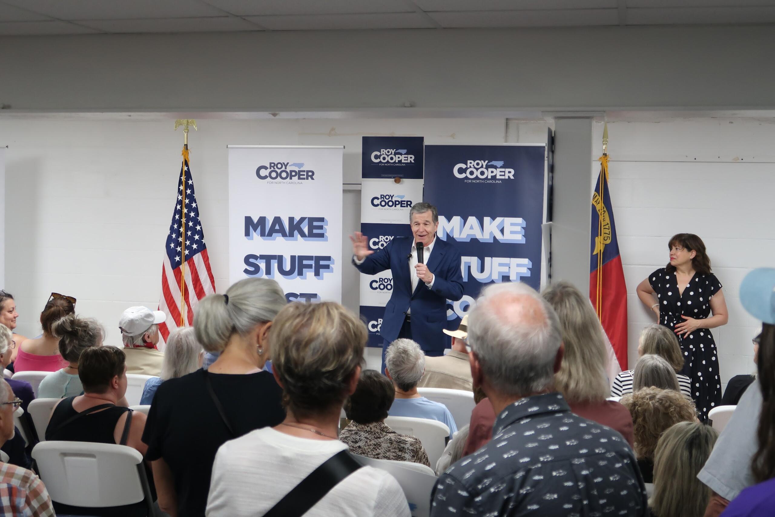 Speaker in a blue suit addresses an indoor political rally, with Roy Cooper banners and American flags behind him.