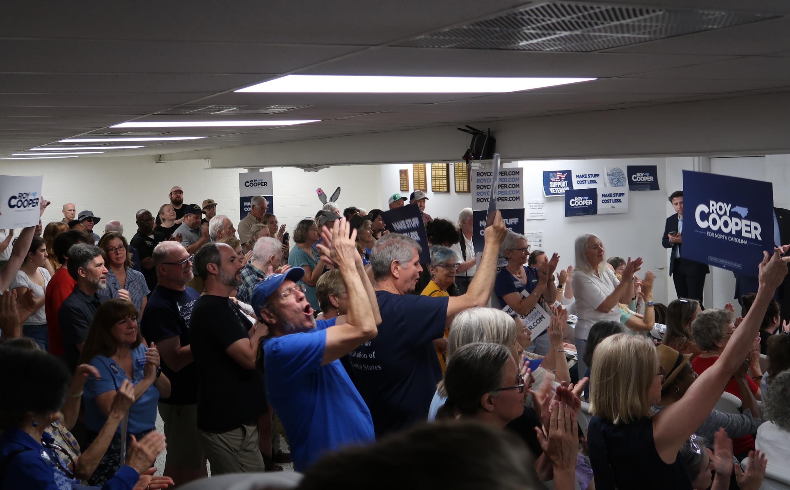 Crowd of supporters at an indoor rally, many with hands raised, holding Roy Cooper campaign signs.