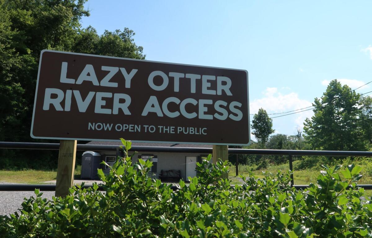 Sign reading 'LAZY OTTER RIVER ACCESS' announcing it is now open to the public, with blue sky and trees in the background.