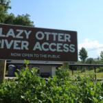 Sign reading 'LAZY OTTER RIVER ACCESS' announcing it is now open to the public, with blue sky and trees in the background.