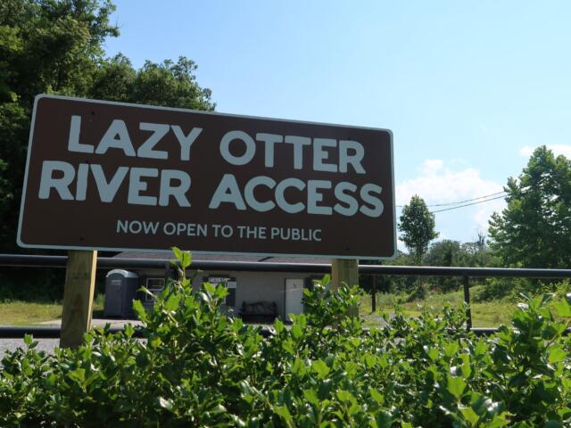 Sign reading 'LAZY OTTER RIVER ACCESS' announcing it is now open to the public, with blue sky and trees in the background.