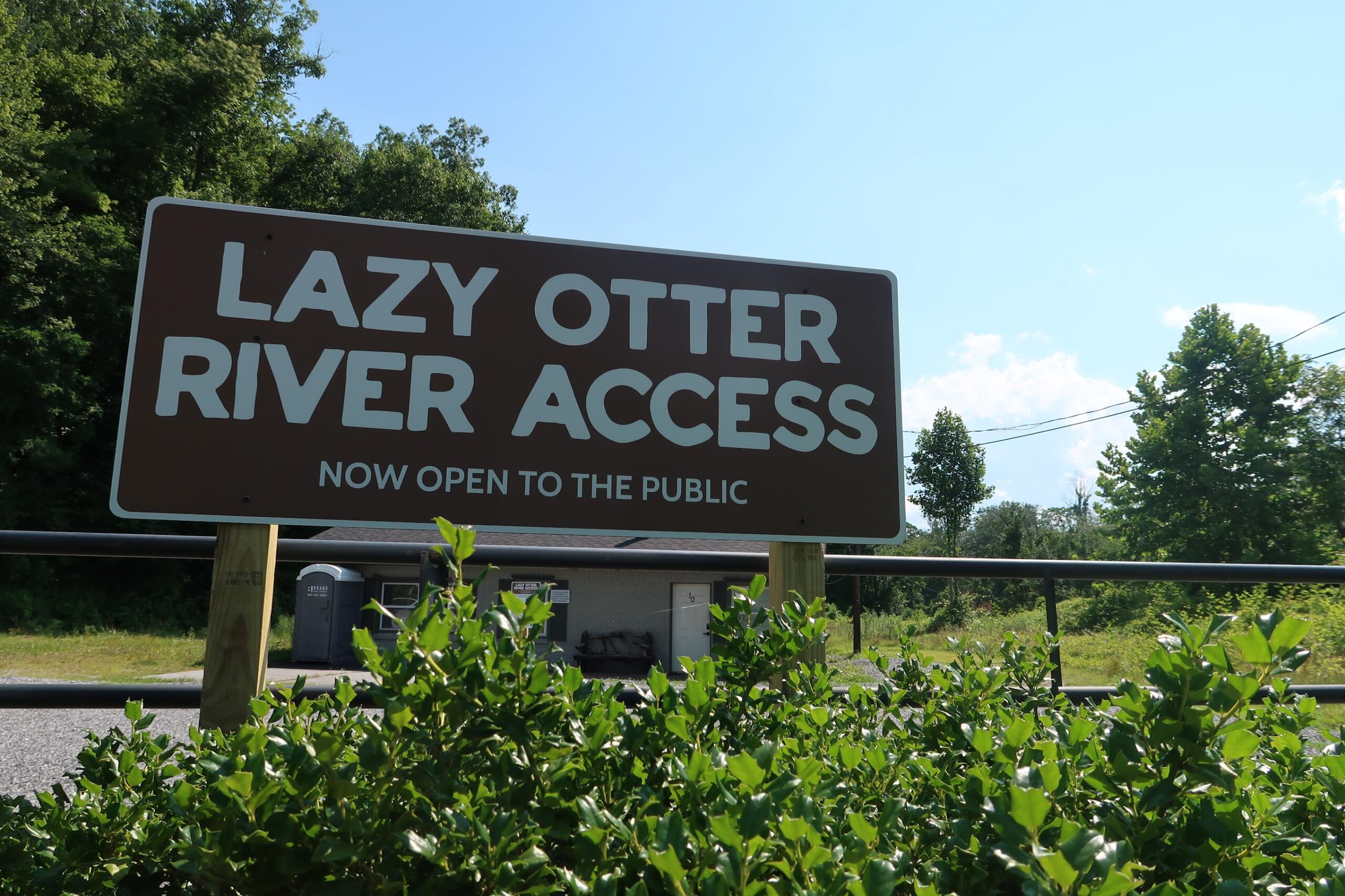 Sign reading 'LAZY OTTER RIVER ACCESS' announcing it is now open to the public, with blue sky and trees in the background.