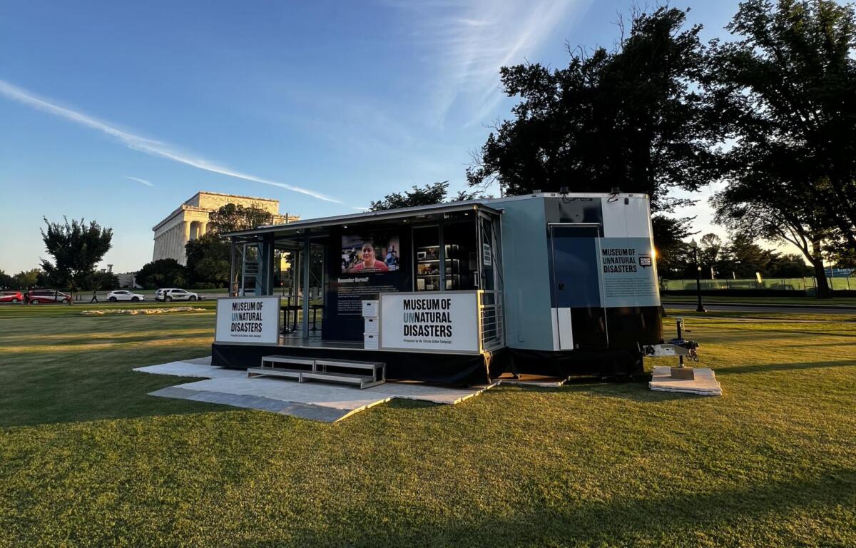 Mobile exhibit trailer labeled Museum of Unnatural Disasters sits on a grassy park with trees and a distant neoclassical building in the background.