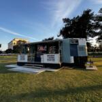 Mobile exhibit trailer labeled Museum of Unnatural Disasters sits on a grassy park with trees and a distant neoclassical building in the background.