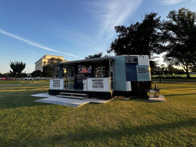 Mobile exhibit trailer labeled Museum of Unnatural Disasters sits on a grassy park with trees and a distant neoclassical building in the background.