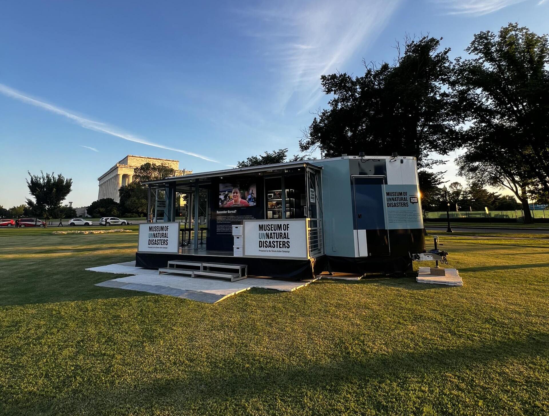 Mobile exhibit trailer labeled Museum of Unnatural Disasters sits on a grassy park with trees and a distant neoclassical building in the background.