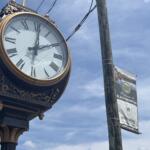 Ornate vintage clock with Roman numerals and gold trim mounted on a decorative post against a blue sky.