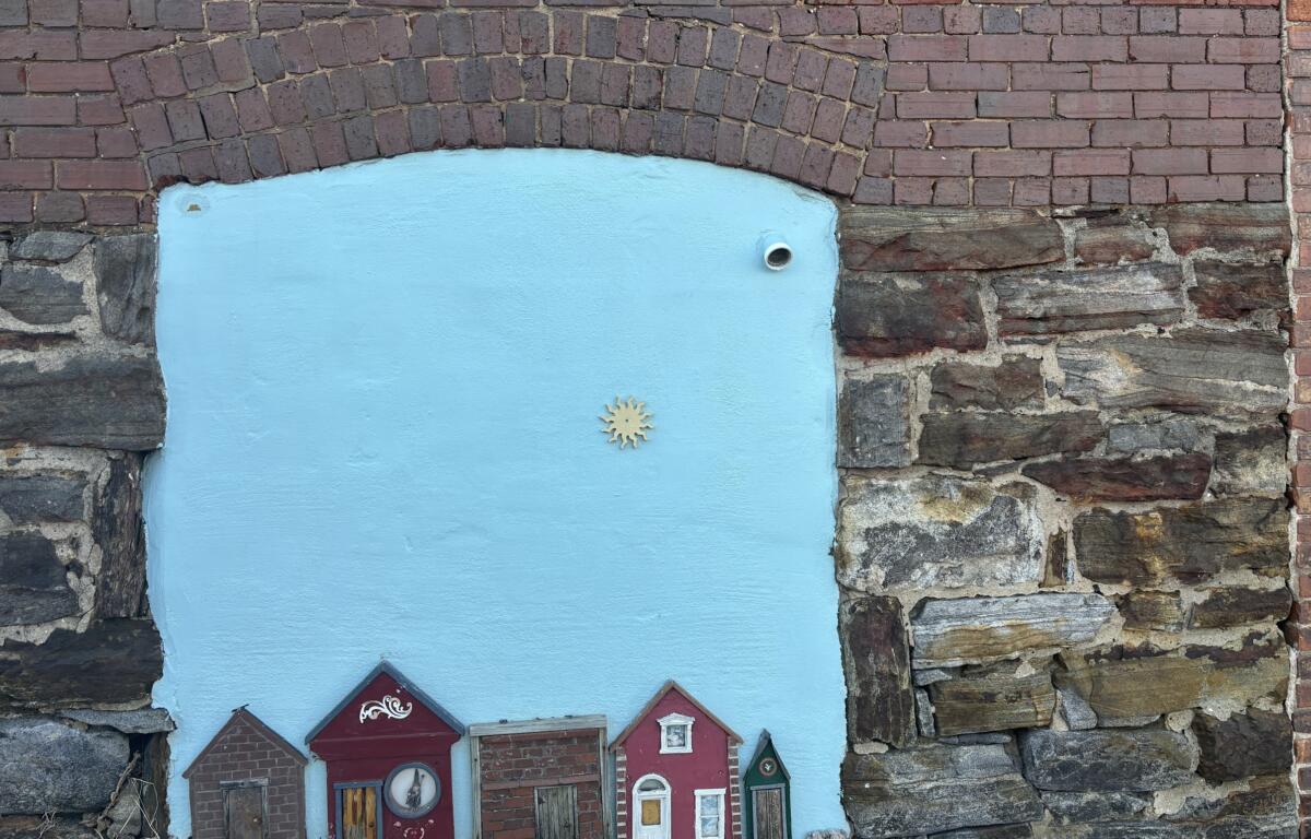 Blue-painted panel set into a brick wall, featuring a row of cute red miniature houses along the bottom and a small sun emblem in the center.