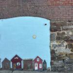 Blue-painted panel set into a brick wall, featuring a row of cute red miniature houses along the bottom and a small sun emblem in the center.