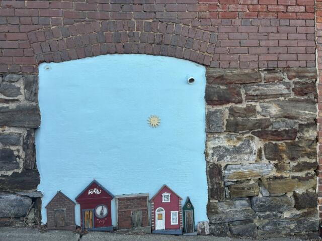 Blue-painted panel set into a brick wall, featuring a row of cute red miniature houses along the bottom and a small sun emblem in the center.