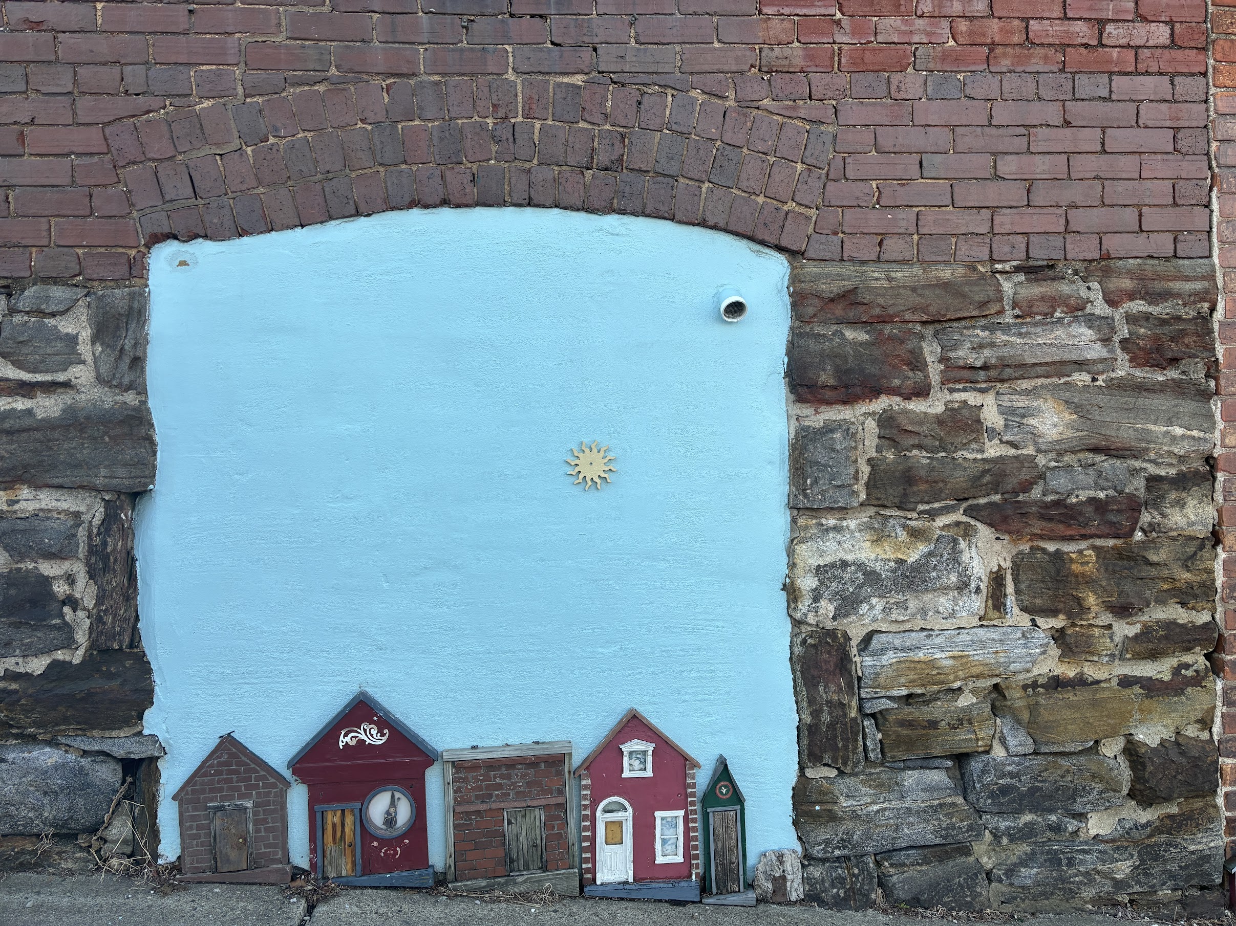 Blue-painted panel set into a brick wall, featuring a row of cute red miniature houses along the bottom and a small sun emblem in the center.
