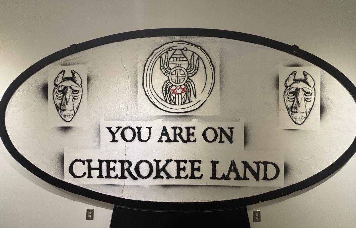 Circular sign reading 'You are on Cherokee land' with two carved mask illustrations on each side of the sign central emblem.