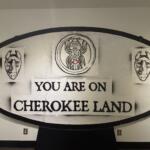 Circular sign reading 'You are on Cherokee land' with two carved mask illustrations on each side of the sign central emblem.