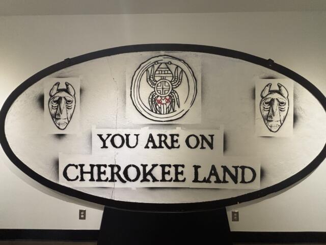 Circular sign reading 'You are on Cherokee land' with two carved mask illustrations on each side of the sign central emblem.
