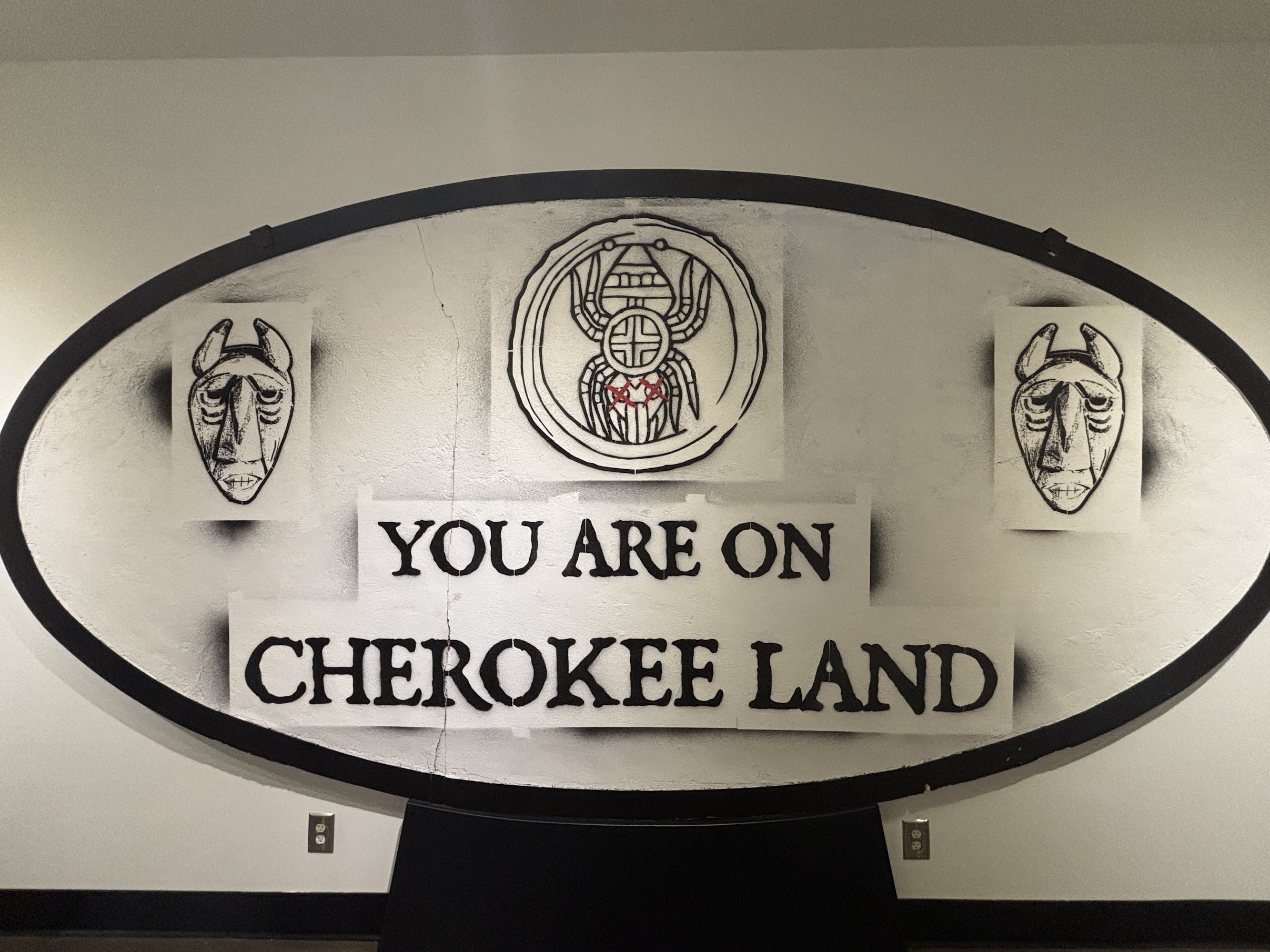 Circular sign reading 'You are on Cherokee land' with two carved mask illustrations on each side of the sign central emblem.