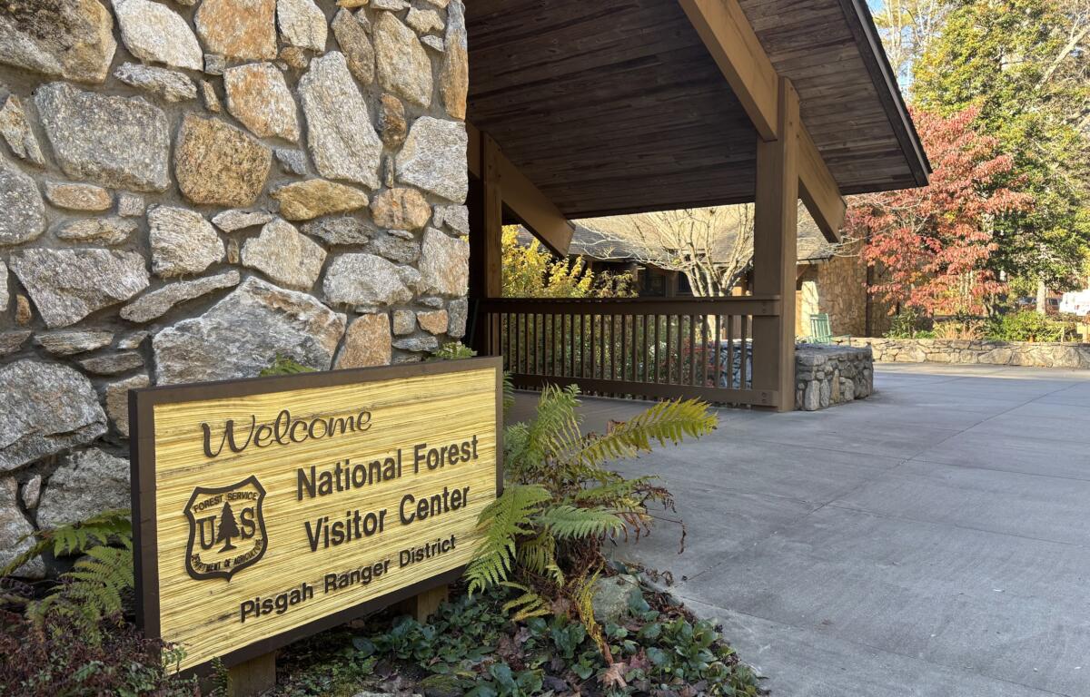 Stone wall with a wooden welcome sign beside a shaded porch of a national forest visitor center, autumn trees in the background.