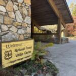 Stone wall with a wooden welcome sign beside a shaded porch of a national forest visitor center, autumn trees in the background.
