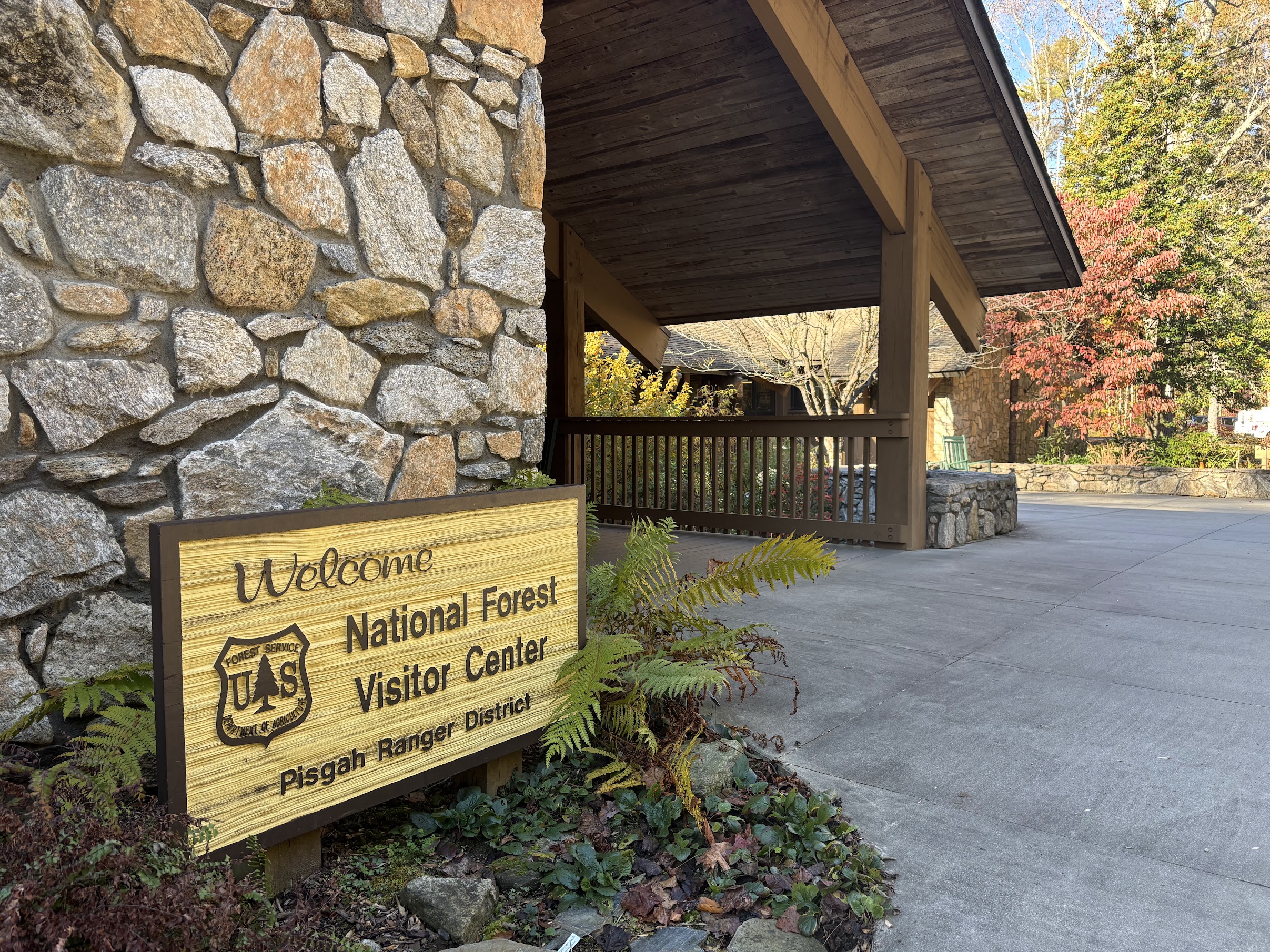 Stone wall with a wooden welcome sign beside a shaded porch of a national forest visitor center, autumn trees in the background.