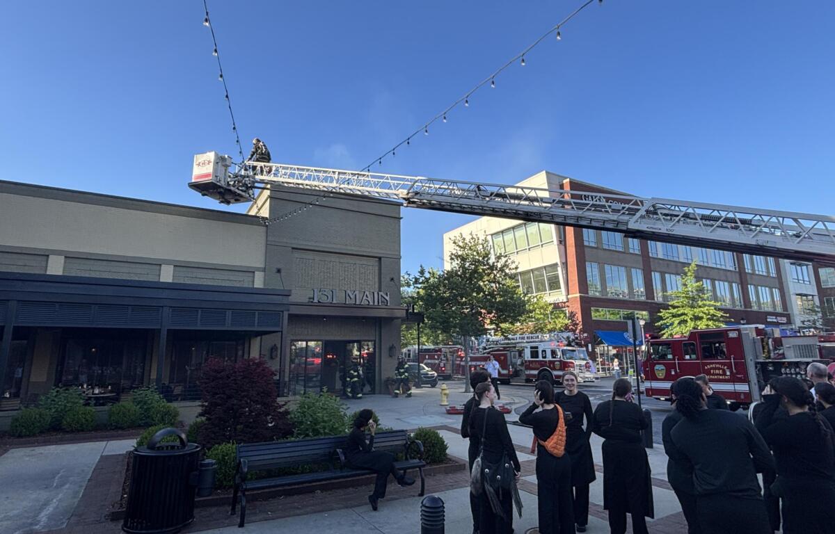 Firefighters on an aerial ladder in front of a brick building, with onlookers and two red fire trucks nearby.