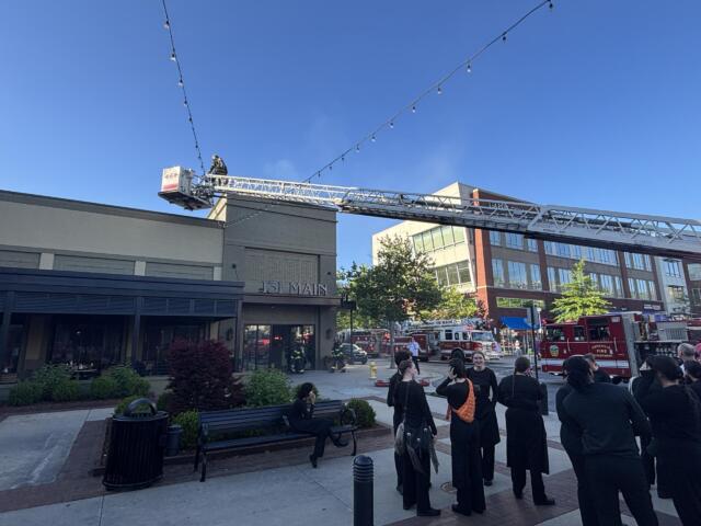Firefighters on an aerial ladder in front of a brick building, with onlookers and two red fire trucks nearby.