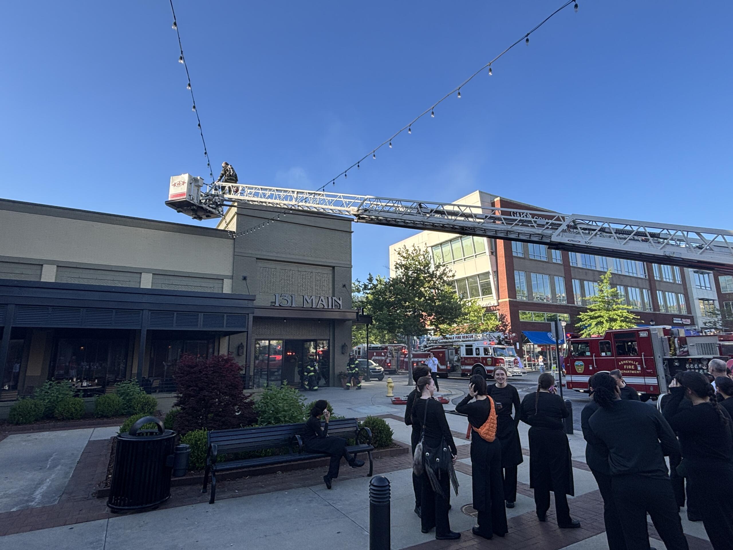 Firefighters on an aerial ladder in front of a brick building, with onlookers and two red fire trucks nearby.