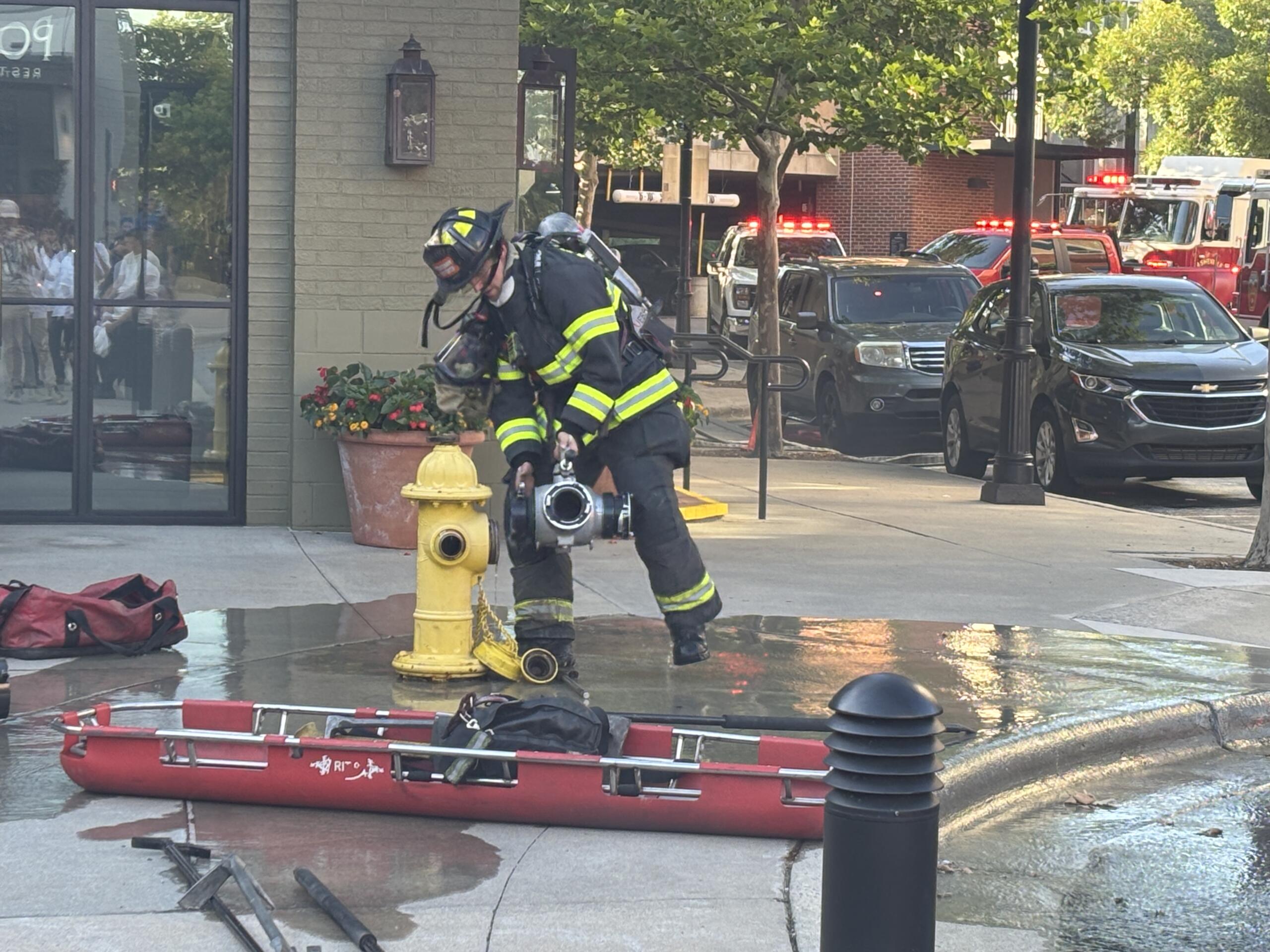 Firefighter in turnout gear handling a hose by a yellow fire hydrant on a sidewalk, with flashing emergency lights behind.