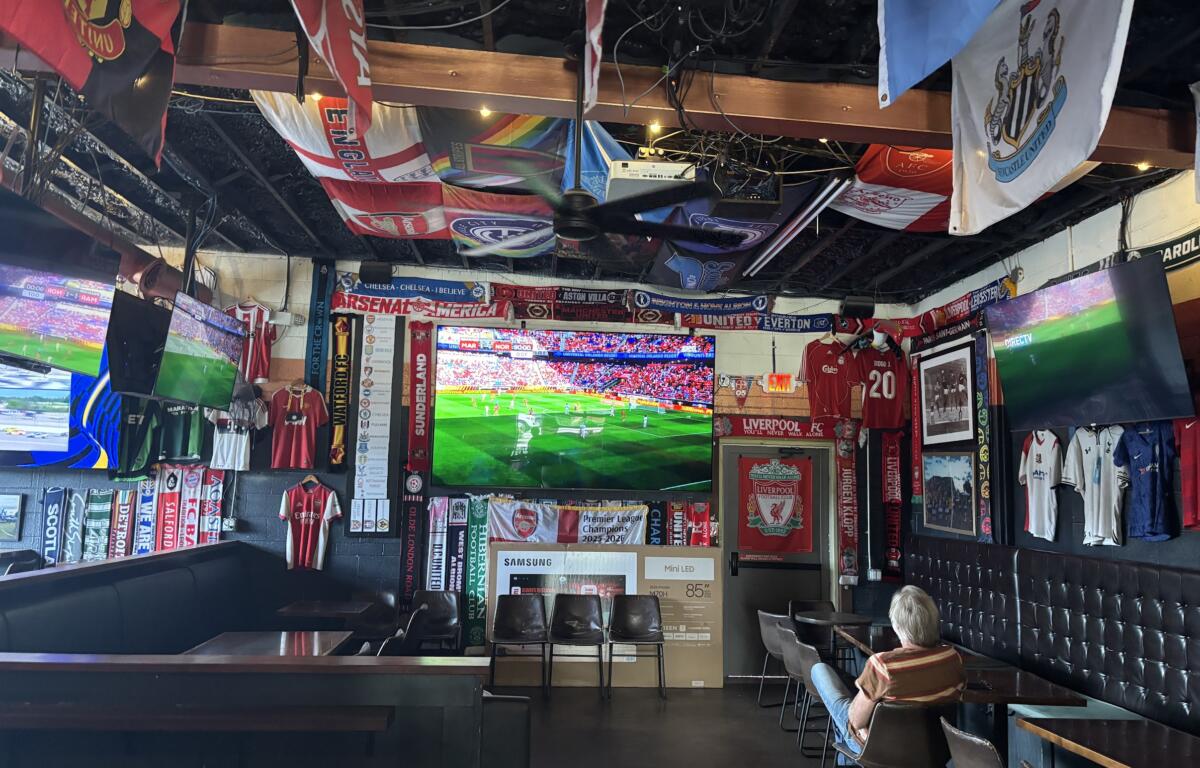 Sports bar interior filled with football banners, flags, and jerseys hanging from the ceiling, with large TV screens showing a live match.