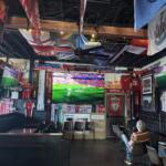 Sports bar interior filled with football banners, flags, and jerseys hanging from the ceiling, with large TV screens showing a live match.