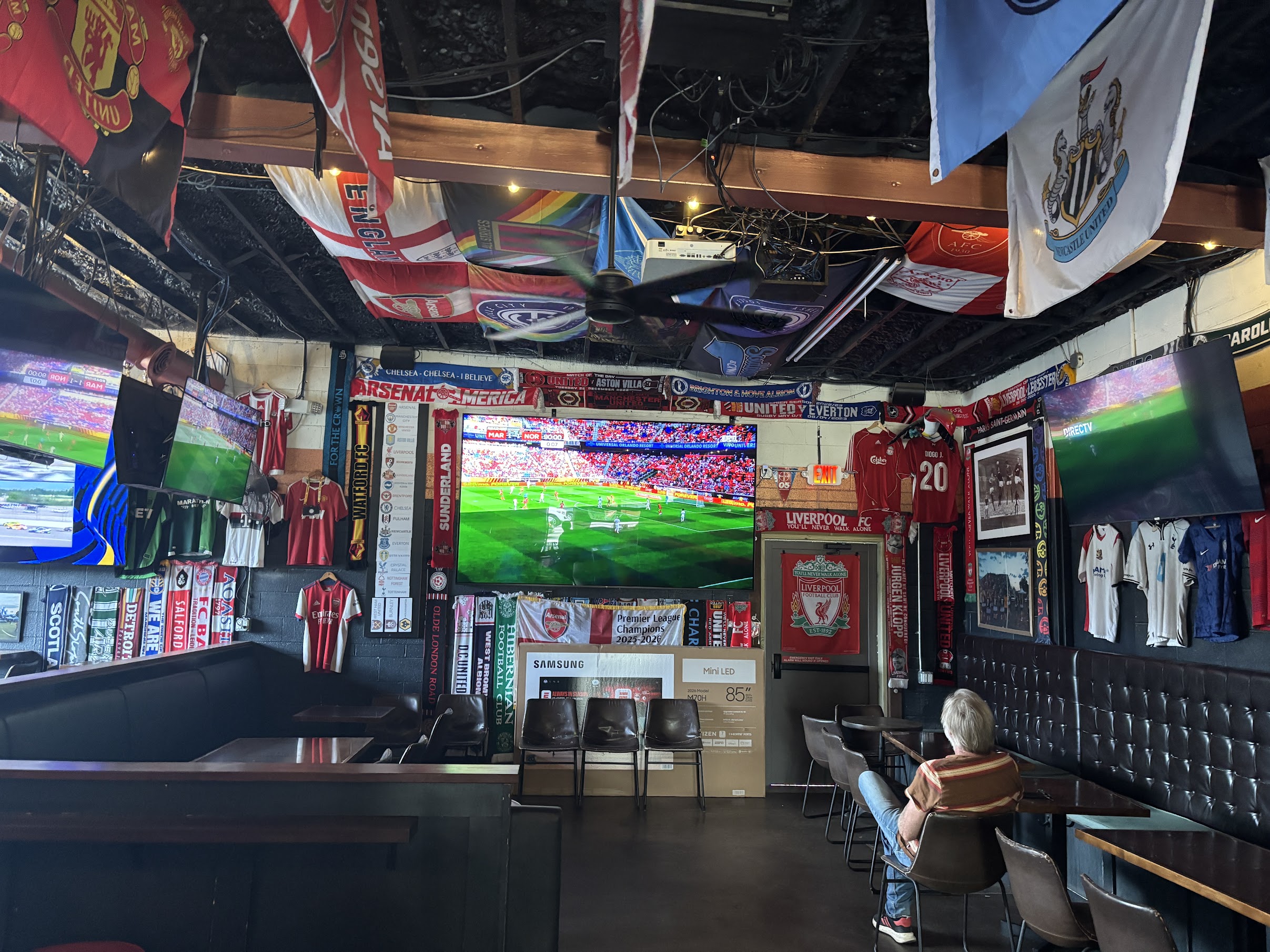 Sports bar interior filled with football banners, flags, and jerseys hanging from the ceiling, with large TV screens showing a live match.