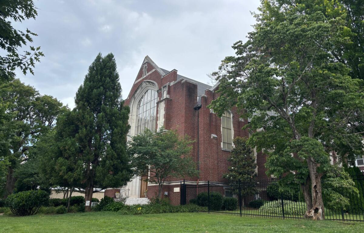 Red brick church with a large arched window, partially obscured by trees on a grassy lawn.