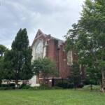 Red brick church with a large arched window, partially obscured by trees on a grassy lawn.