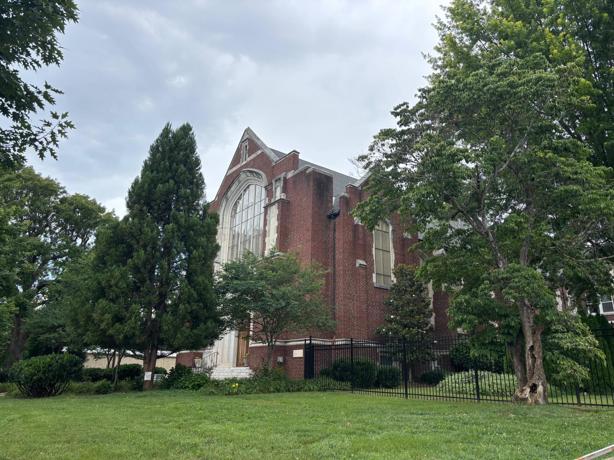 Red brick church with a large arched window, partially obscured by trees on a grassy lawn.