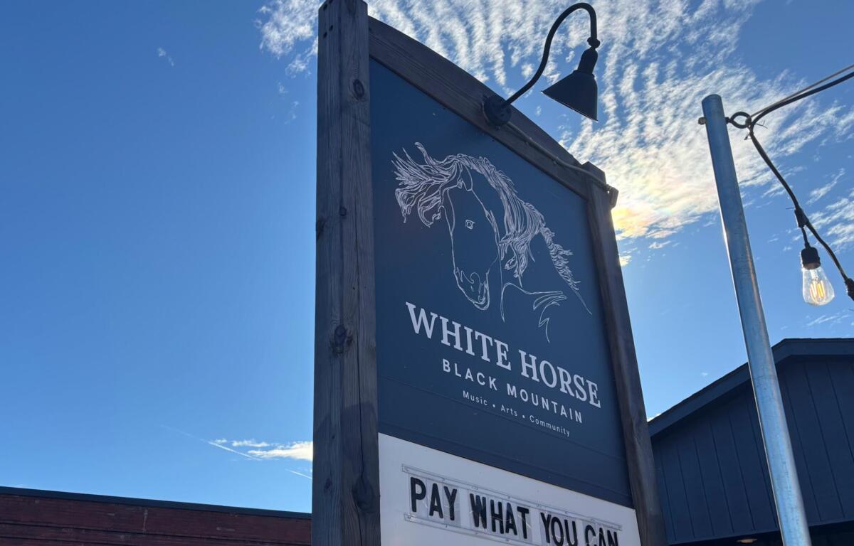 Wooden-framed sign under a blue sky with a white horse illustration and the words 'White Horse Black Mountain' and a 'Pay What You Can' panel nearby.