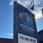 Wooden-framed sign under a blue sky with a white horse illustration and the words 'White Horse Black Mountain' and a 'Pay What You Can' panel nearby.