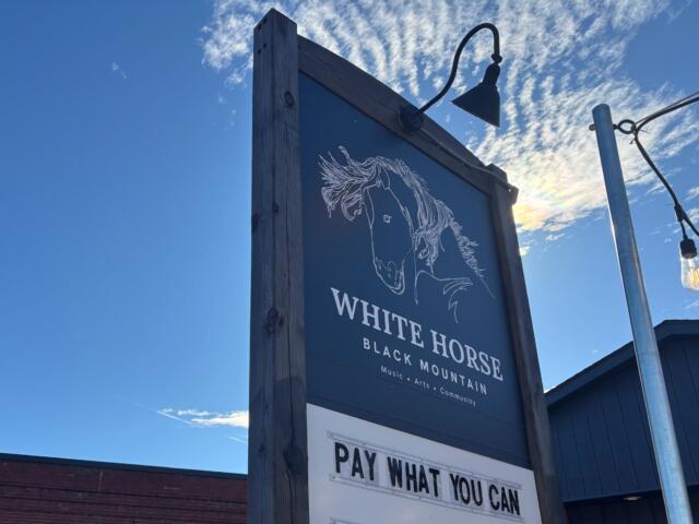 Wooden-framed sign under a blue sky with a white horse illustration and the words 'White Horse Black Mountain' and a 'Pay What You Can' panel nearby.