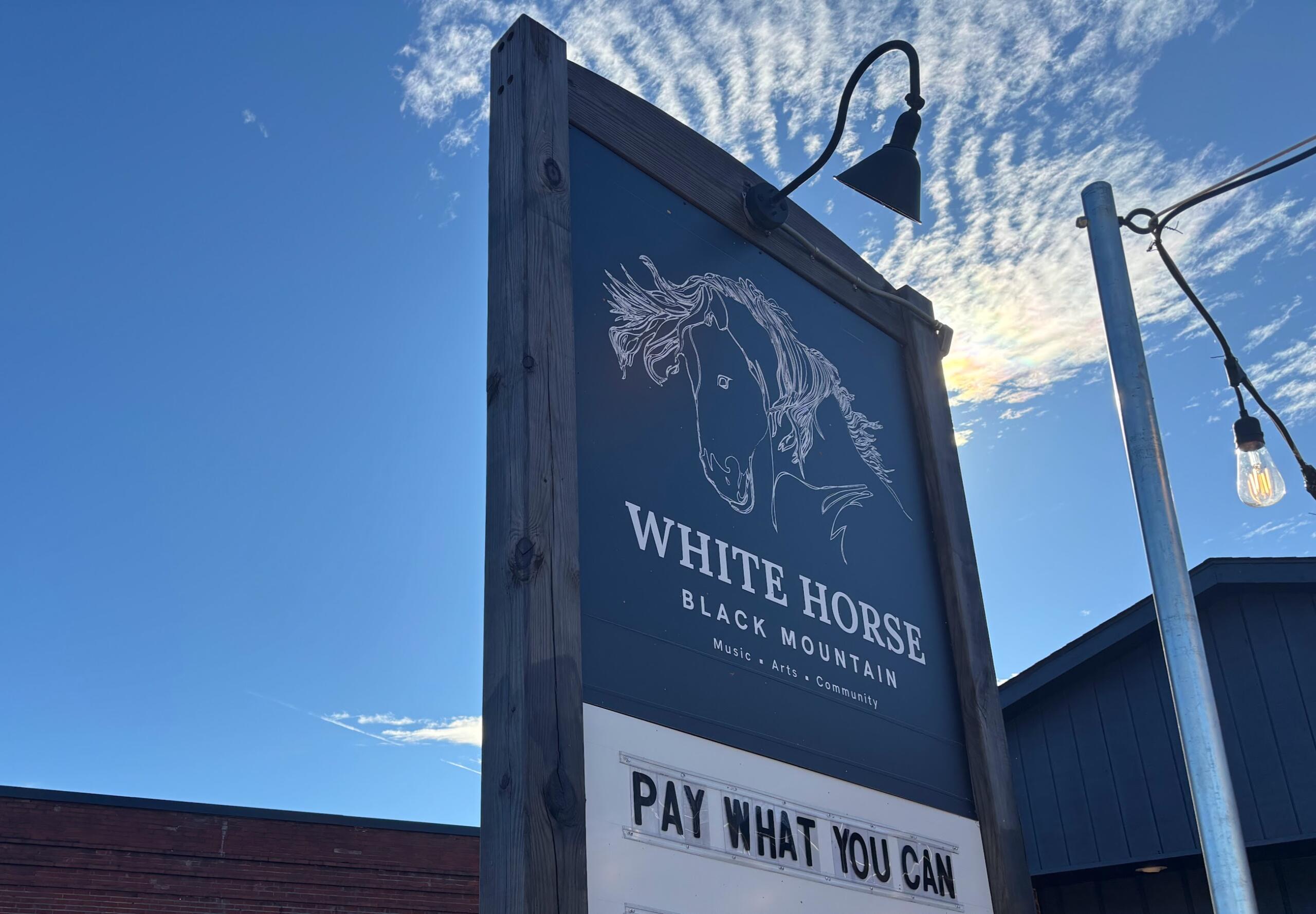 Wooden-framed sign under a blue sky with a white horse illustration and the words 'White Horse Black Mountain' and a 'Pay What You Can' panel nearby.