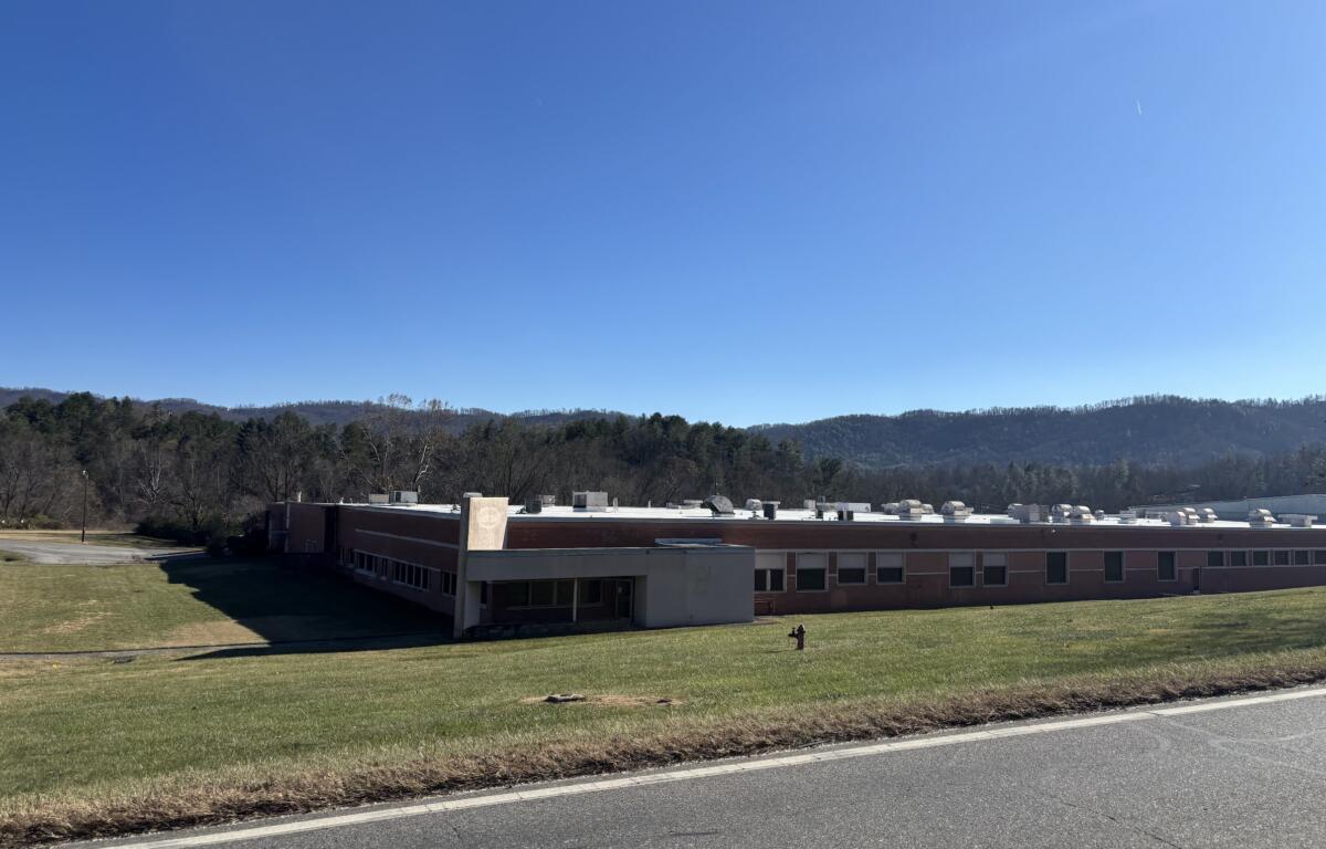 Single-story brick school building with a flat roof and a grassy front lawn, set against a clear blue sky and tree-covered hills in the distance.