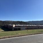 Single-story brick school building with a flat roof and a grassy front lawn, set against a clear blue sky and tree-covered hills in the distance.
