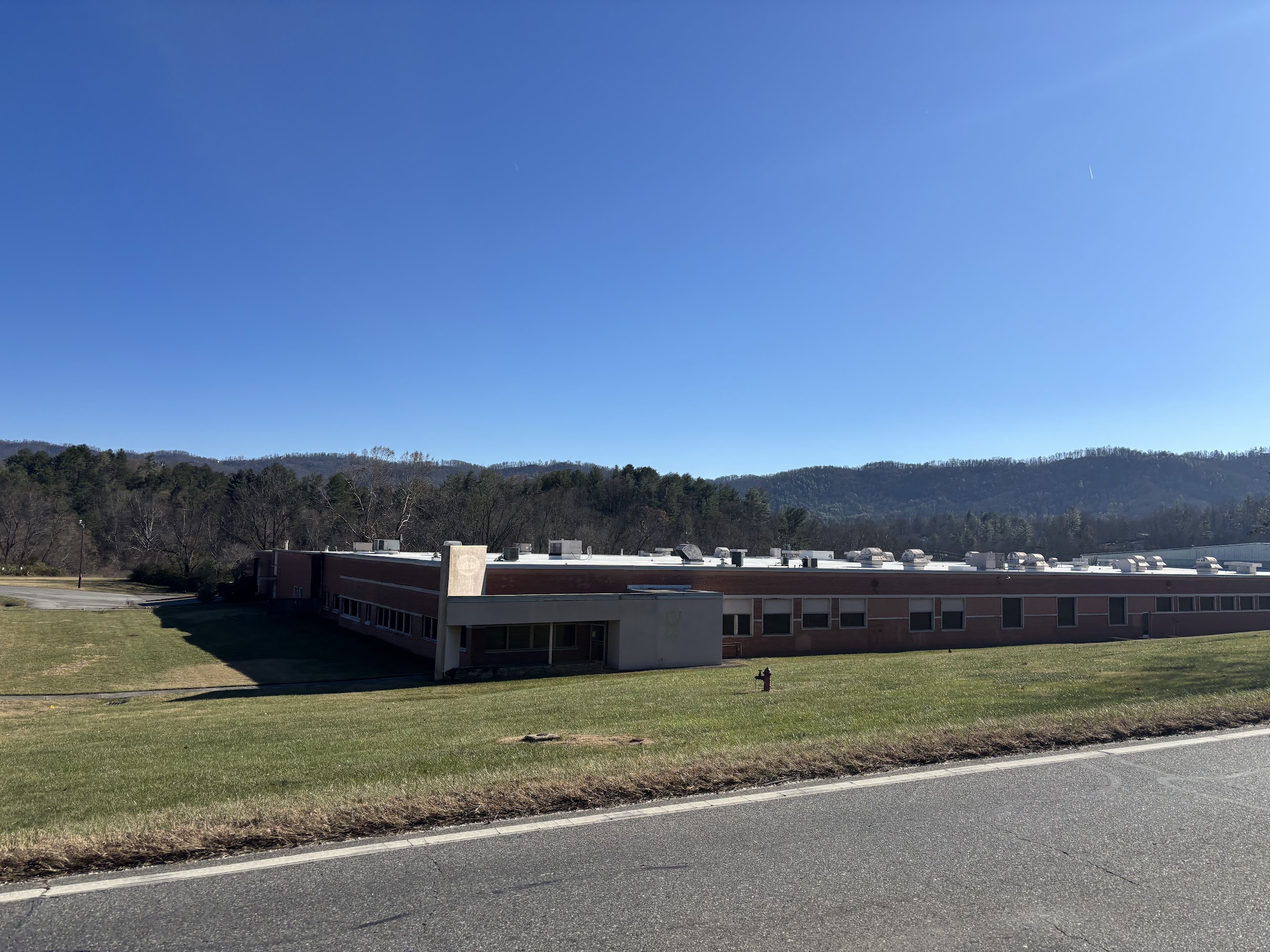 Single-story brick school building with a flat roof and a grassy front lawn, set against a clear blue sky and tree-covered hills in the distance.