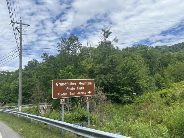 Brown road sign reading 'Grandfather Mountain State Park' with arrows and 'Profile Trail Access' in a wooded roadside area, beside a guardrail and power lines.