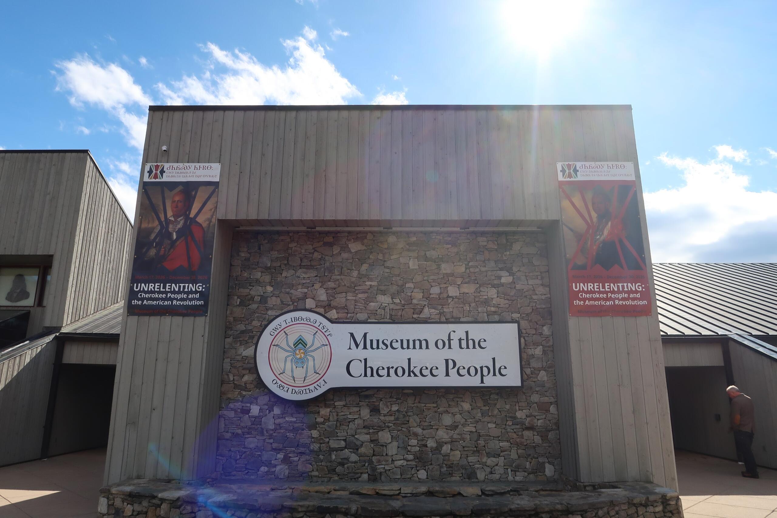 Front view of a museum building with stone wall and banners advertising the Cherokee exhibit under a bright sky.