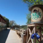 Exterior view of a nature center: a tall wooden sign with a circular logo and 'Nature Center' text, beside a mural-painted wall and visitors on a sunny walkway.