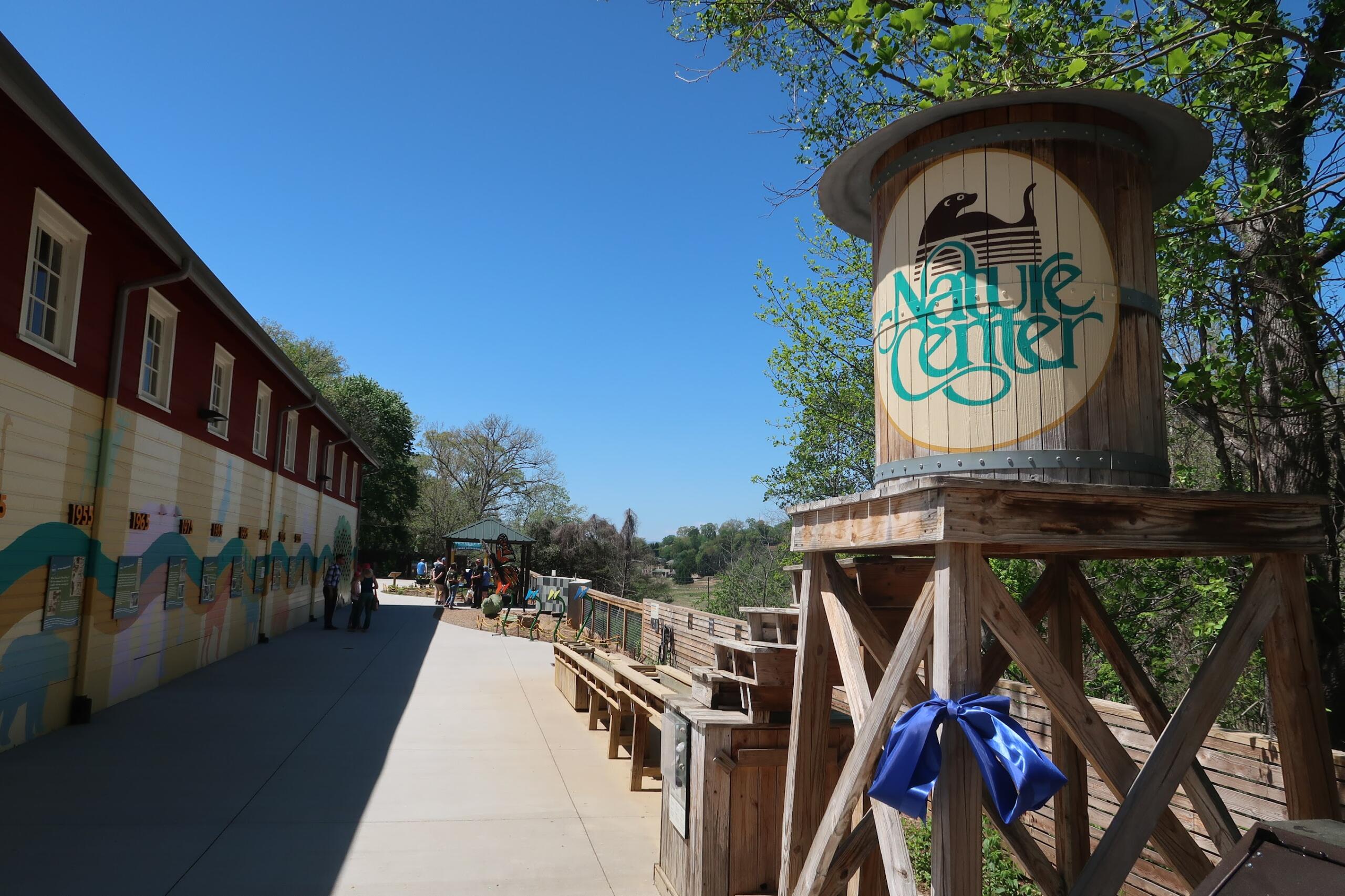Exterior view of a nature center: a tall wooden sign with a circular logo and 'Nature Center' text, beside a mural-painted wall and visitors on a sunny walkway.