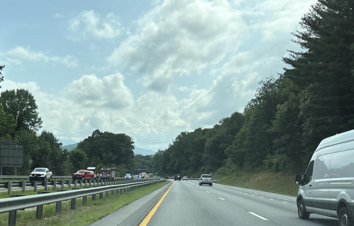 Highway scene with cars in both directions, guardrails, and forested hills under a partly cloudy sky.