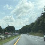Highway scene with cars in both directions, guardrails, and forested hills under a partly cloudy sky.