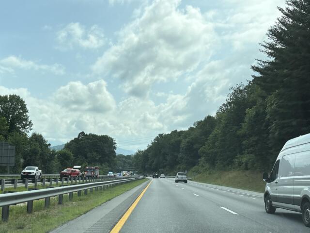 Highway scene with cars in both directions, guardrails, and forested hills under a partly cloudy sky.