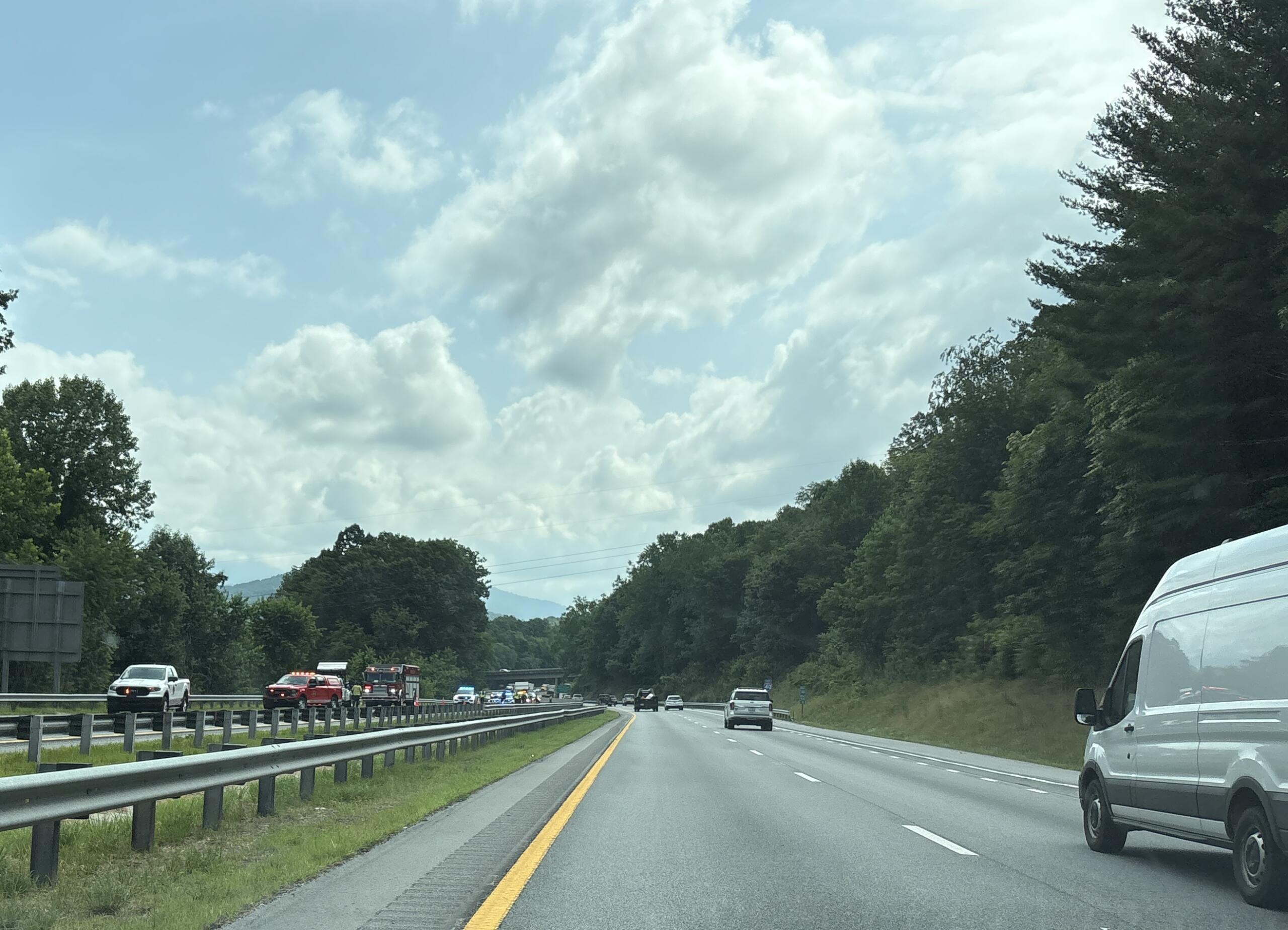 Highway scene with cars in both directions, guardrails, and forested hills under a partly cloudy sky.