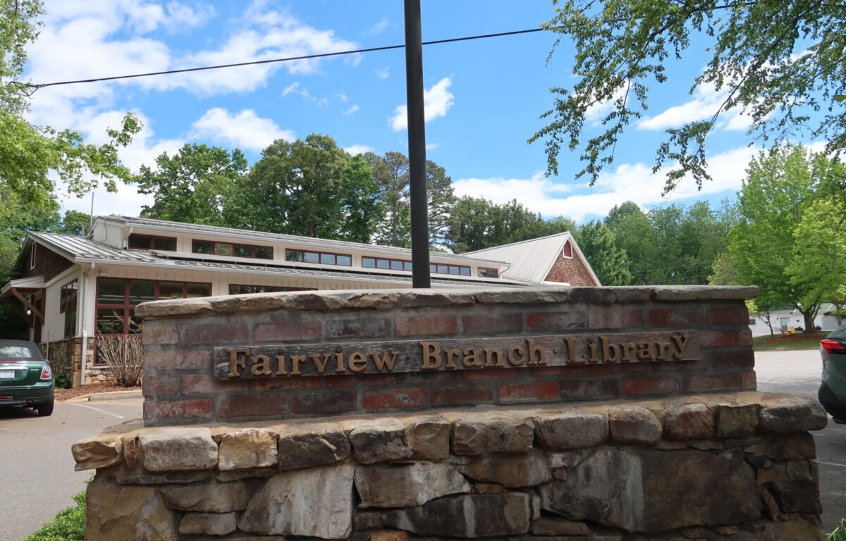 Stone brick sign wall with carved text reading 'Fairview Branch Library' in front of a low building and green trees under a blue sky.